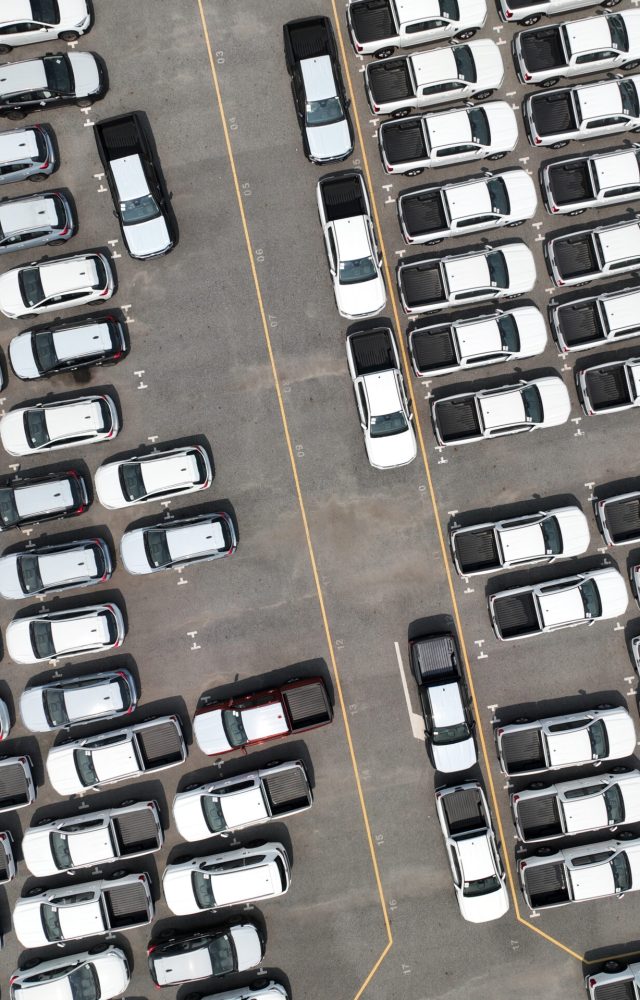 Aerial view of new cars stock at factory parking lot. Above view cars parked in a row. Automotive industry. Logistics business. Import or export new cars at warehouse. Big parking lot at port terminal